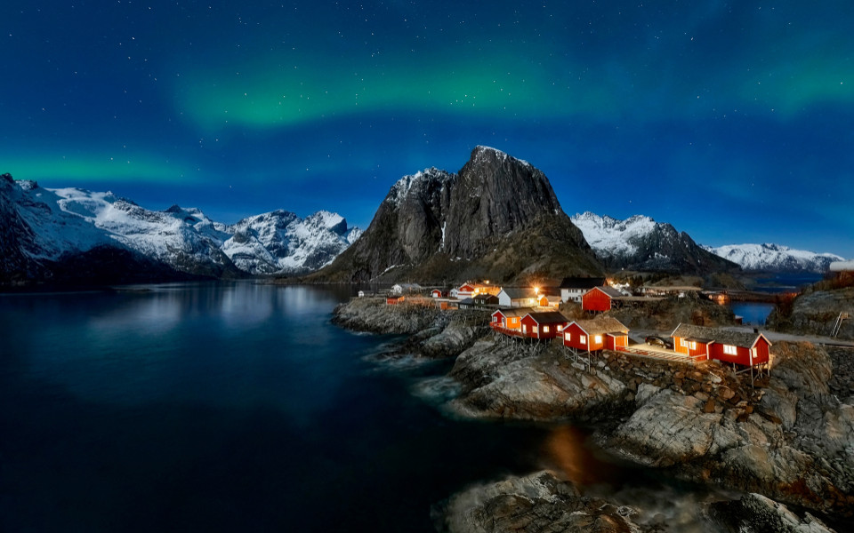 Nordlichter &uuml;ber roten Fischerh&uuml;tten in einem norwegischen Fjord, umgeben von schneebedeckten Bergen bei Nacht.