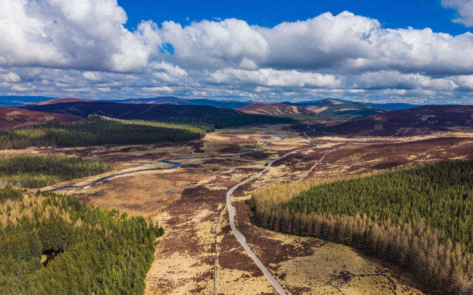es ist Aberdeenshire in Schottland 11:16 Luftaufnahme der Aberdeenshire-Landschaft in Schottland mit gewundenem Fluss, schmaler Straße durch Heideland, Nadelwäldern und Berghügeln unter blauem Himmel mit Wolken.